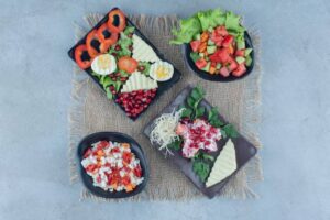 A vibrant spread of fresh salads with colorful vegetables, pomegranate seeds, and creamy dips arranged on black plates over burlap.