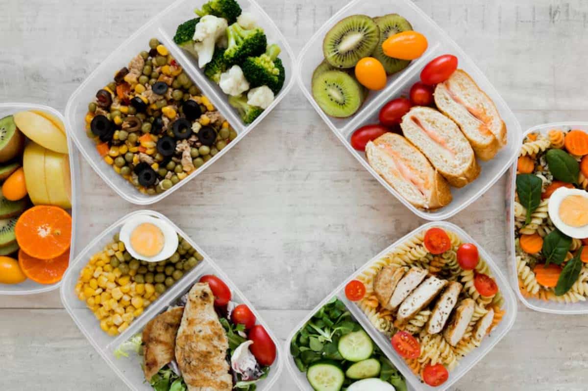 A variety of healthy meal prep containers filled with fruits, vegetables, proteins, and grains, arranged on a light wooden surface.