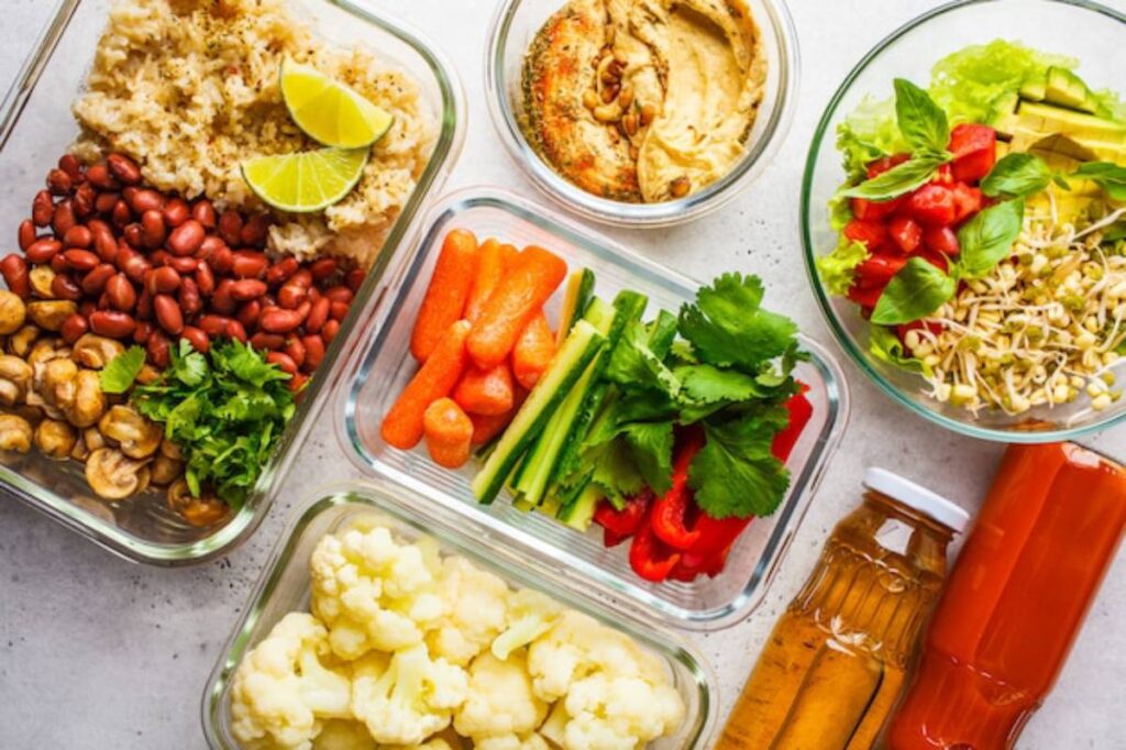 Colorful meal prep containers filled with grains, beans, fresh vegetables, herbs, hummus, and drinks on a light background.