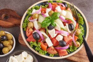 A vibrant salad featuring lettuce, tomatoes, mozzarella, olives, onions, and basil, served on a blue plate with olive dishes on the side.