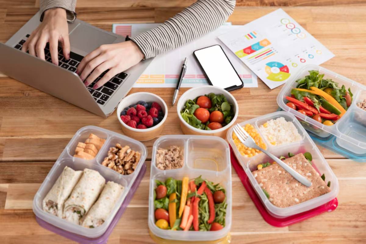 A person types on a laptop surrounded by healthy meal prep containers, fruits, and a smartphone, on a wooden table.