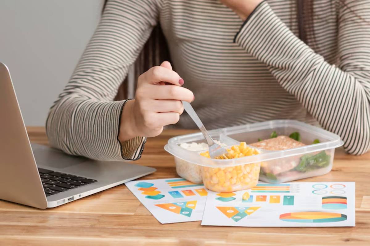 A person in a striped shirt eats from a meal container while working on a laptop, surrounded by colorful charts and graphs.
