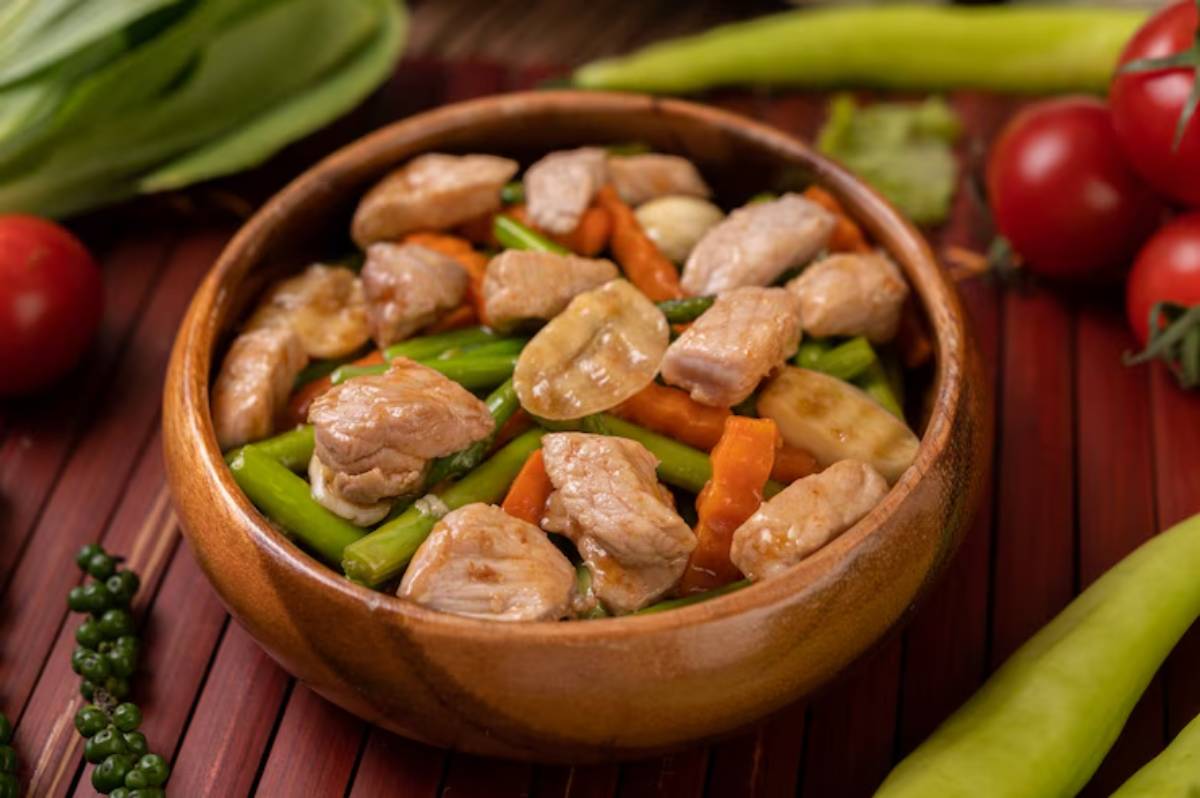 A wooden bowl filled with tender pork pieces and colorful mixed vegetables, including carrots, green beans, and garlic.