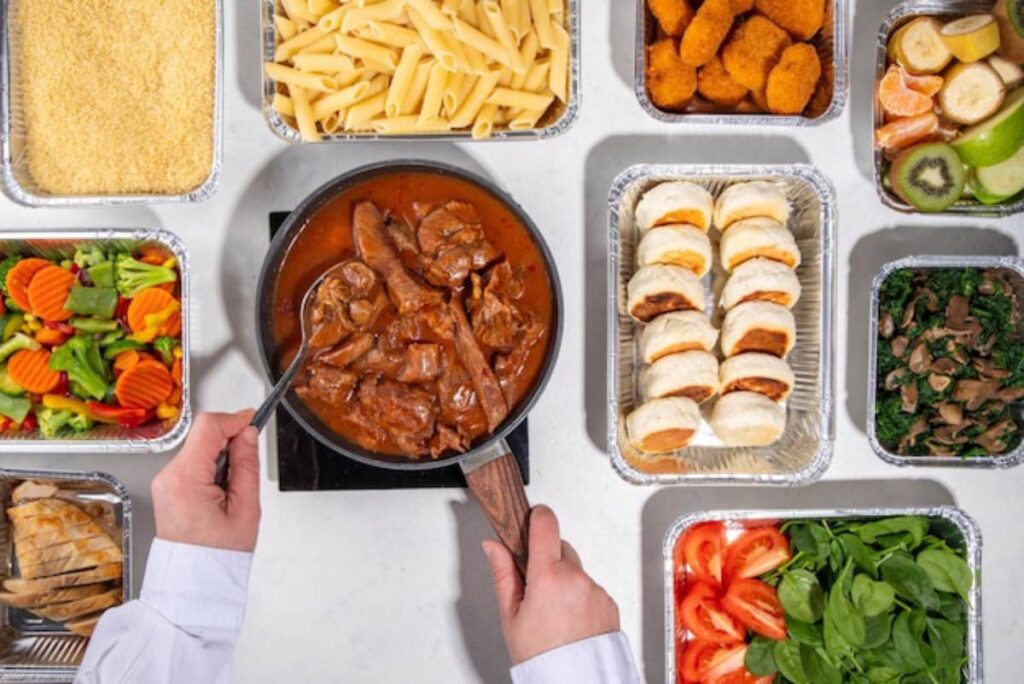 A hand holding a spoon stirs beef in rich sauce, surrounded by various prepared dishes: pasta, salad, chicken, rolls, and fruit.