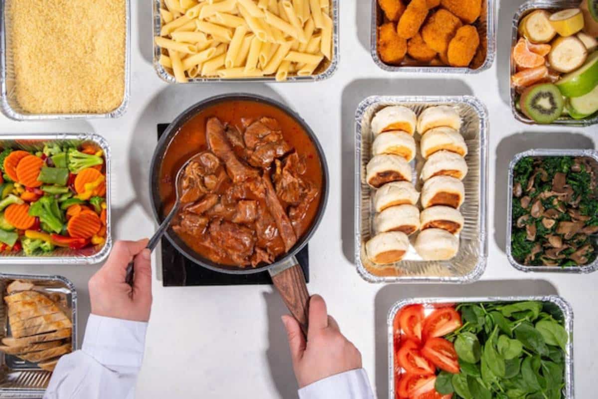 A hand holding a spoon stirs beef in rich sauce, surrounded by various prepared dishes: pasta, salad, chicken, rolls, and fruit.
