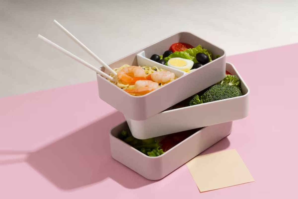 A stack of three modern food containers filled with shrimp noodles, vegetables, salad, and boiled egg, with chopsticks resting on top.