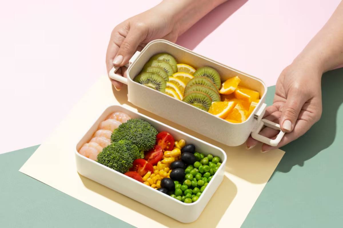 A person holding a bento box with vibrant fruit slices on top and colorful veggies and shrimp in the bottom compartment.