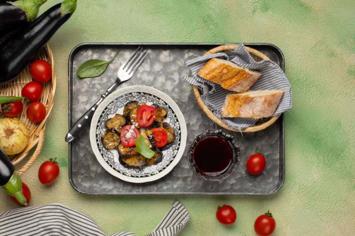 A plate of grilled vegetables with cherry tomatoes, a glass of red wine, and bread rolls on a textured green background.