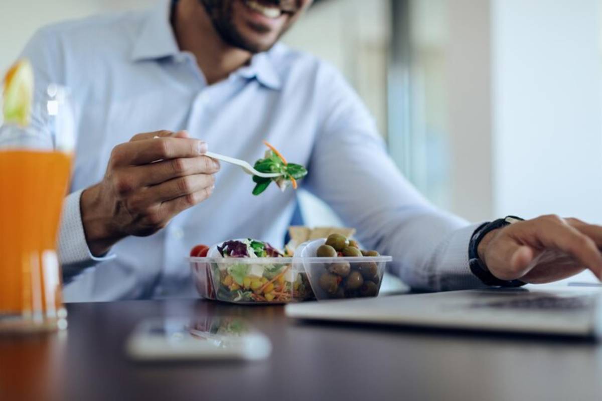 A man in a blue shirt enjoys a healthy lunch of salad and olives while working on a laptop, with a drink beside him.