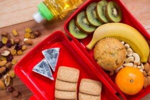 A colorful lunchbox containing sliced kiwi, a banana, muffin, orange, nuts, cookies, and a bottle of oil on a wooden surface.