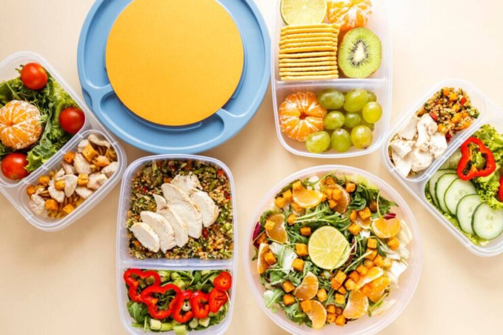 A vibrant display of meal prep containers filled with salads, fruits, and proteins, centered around a large yellow plate.