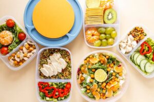 A vibrant display of meal prep containers filled with salads, fruits, and proteins, centered around a large yellow plate.