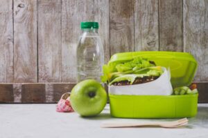 A green lunchbox with a sandwich and veggies, an apple, and a water bottle are placed against a rustic wooden backdrop.