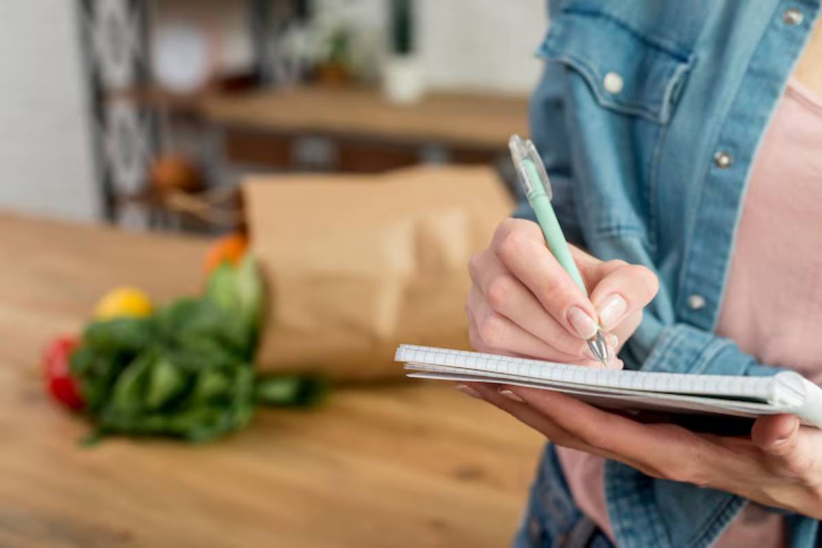A person in a denim jacket writes in a notebook while fresh vegetables are arranged on a wooden table in the background.