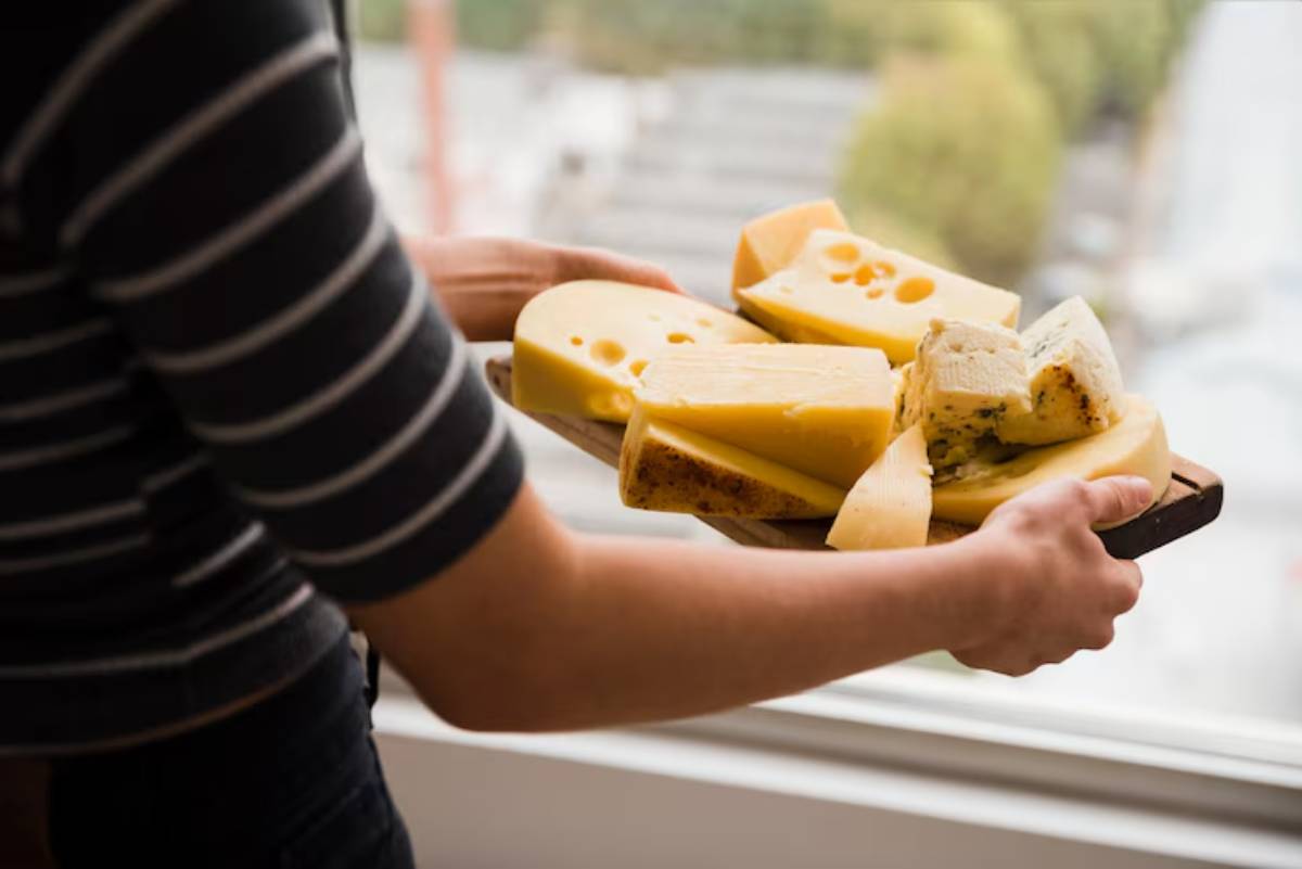 A person holds a wooden platter filled with various types of cheese, with a blurred urban view in the background.