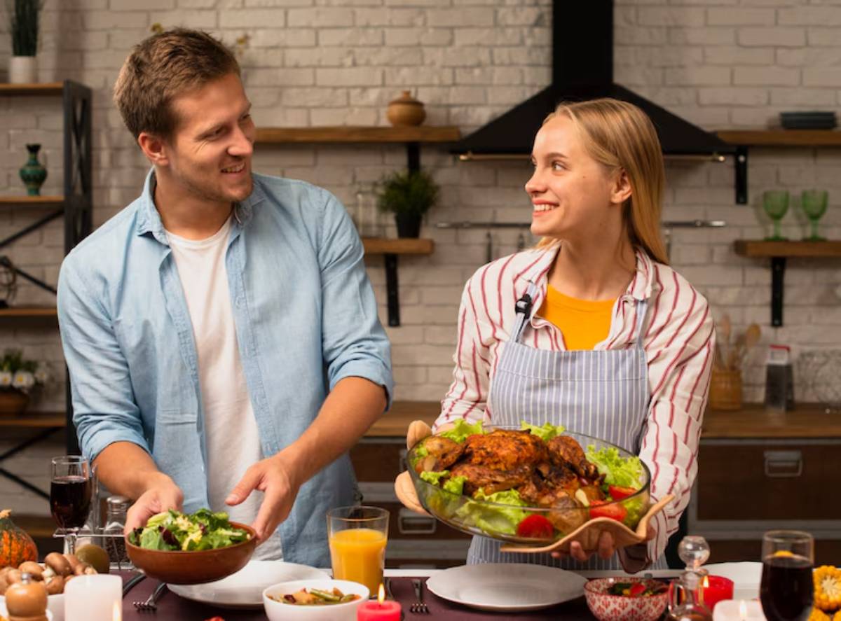 A man and woman proudly present a beautifully arranged meal with salad and roasted chicken on a rustic kitchen table.