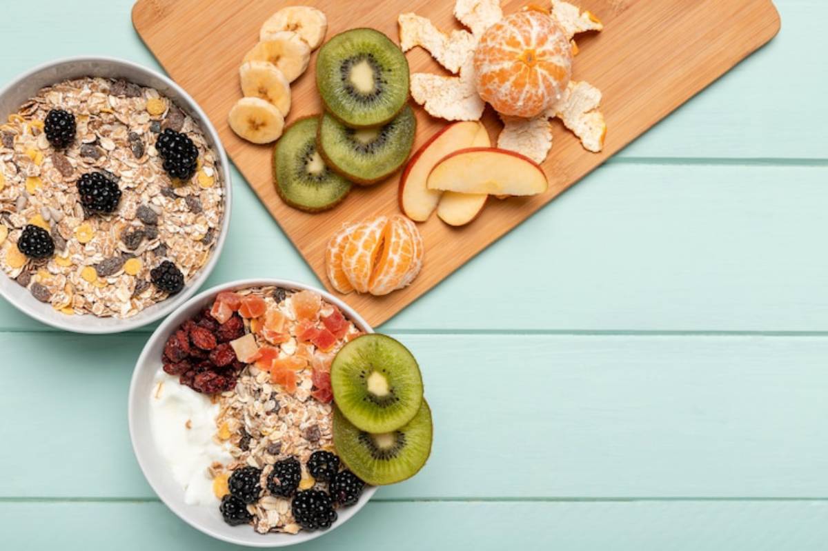 Two bowls of colorful oatmeal topped with fruits, alongside a wooden board of sliced bananas, kiwi, apple, and a mandarin orange.