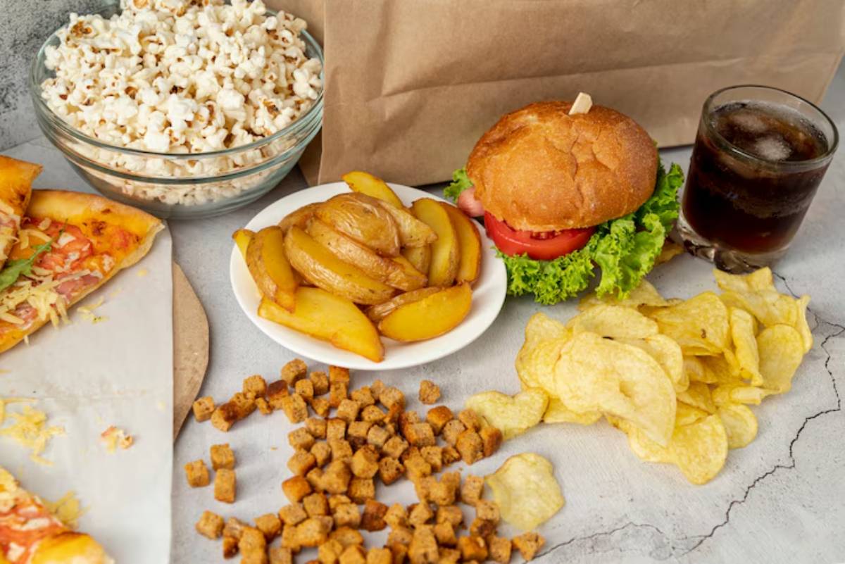 A spread of snacks including popcorn, a burger with lettuce and tomato, baked apple slices, chips, croutons, and pizza slices.