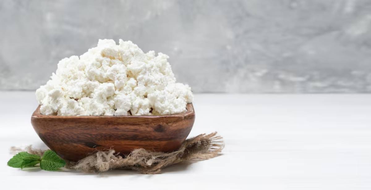 A wooden bowl filled with fluffy, white cottage cheese sits on a rustic surface, accompanied by a sprig of mint.