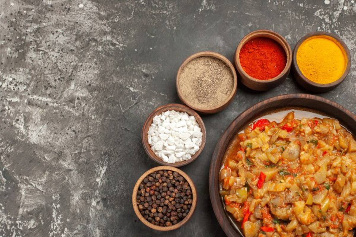 A bowl of colorful vegetable curry surrounded by wooden bowls of spices: black pepper, salt, red chili, and turmeric on a dark countertop.