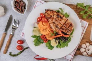 Grilled chicken breast served on a bed of fresh vegetables, garnished with lime, chilies, and herbs, with garlic and dried peppers in the background.