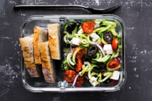 Fresh salad with spiralized zucchini, cherry tomatoes, olives, feta cheese, and slices of whole grain bread in a glass container.
