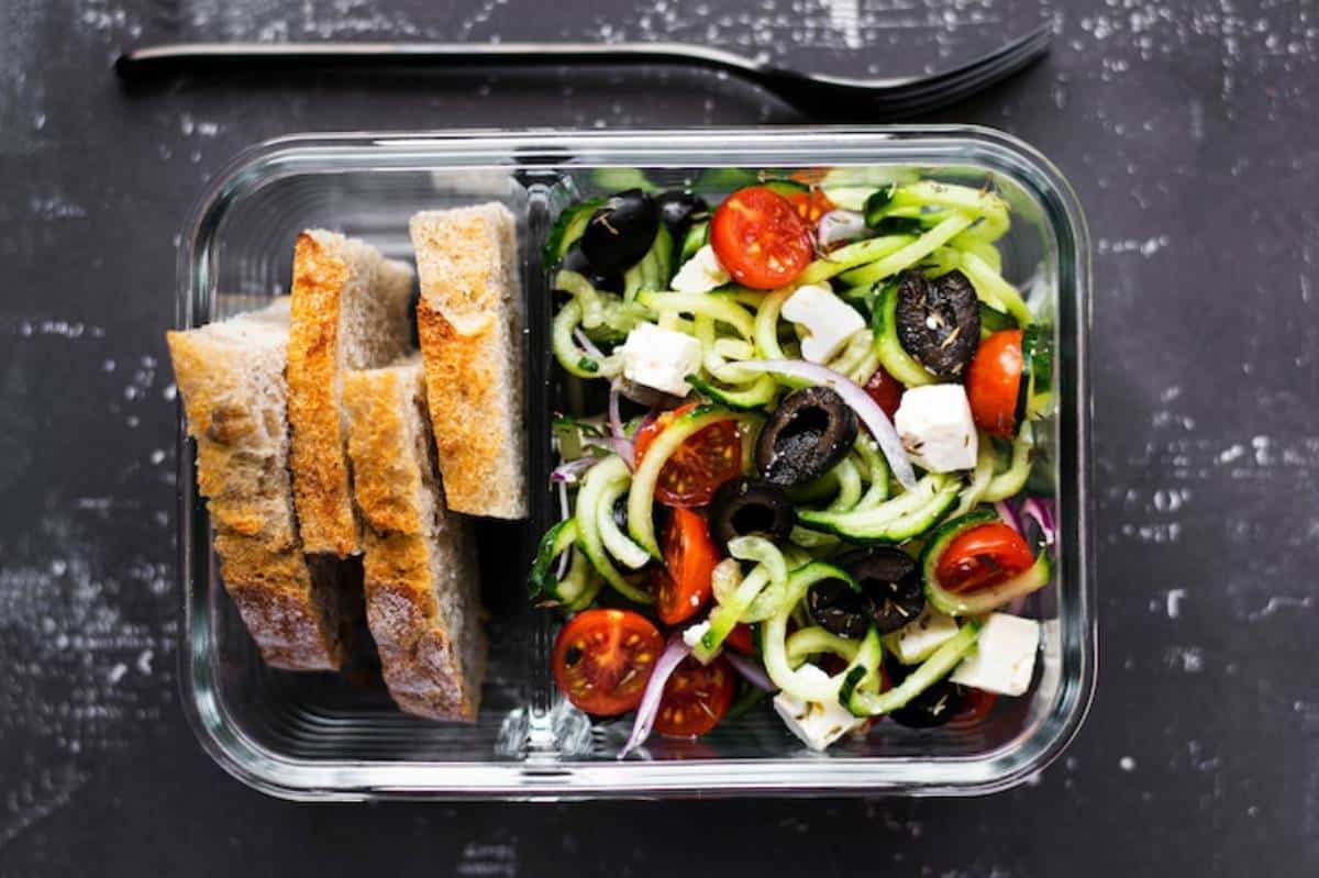 Fresh salad with spiralized zucchini, cherry tomatoes, olives, feta cheese, and slices of whole grain bread in a glass container.