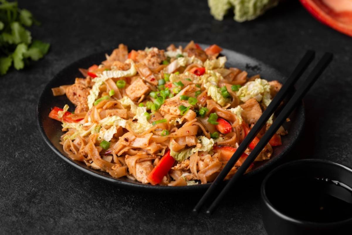 A black plate of stir-fried noodles with chicken, cabbage, and red peppers, garnished with green onions, and chopsticks on the side.