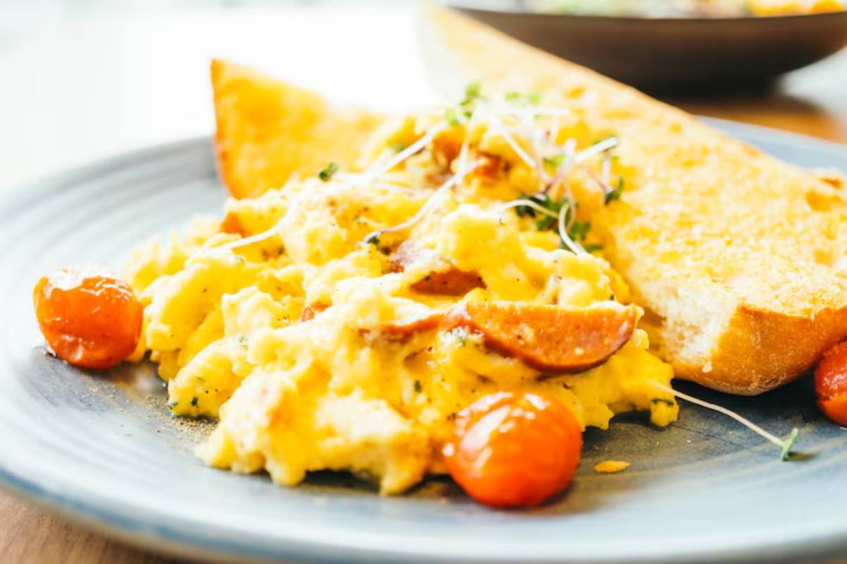 A plate of creamy scrambled eggs with sliced sausage and cherry tomatoes, accompanied by a slice of toasted bread.