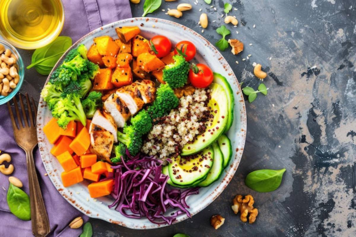 A vibrant bowl featuring grilled chicken, quinoa, broccoli, sweet potatoes, cherry tomatoes, cucumber, and purple cabbage on a rustic surface.