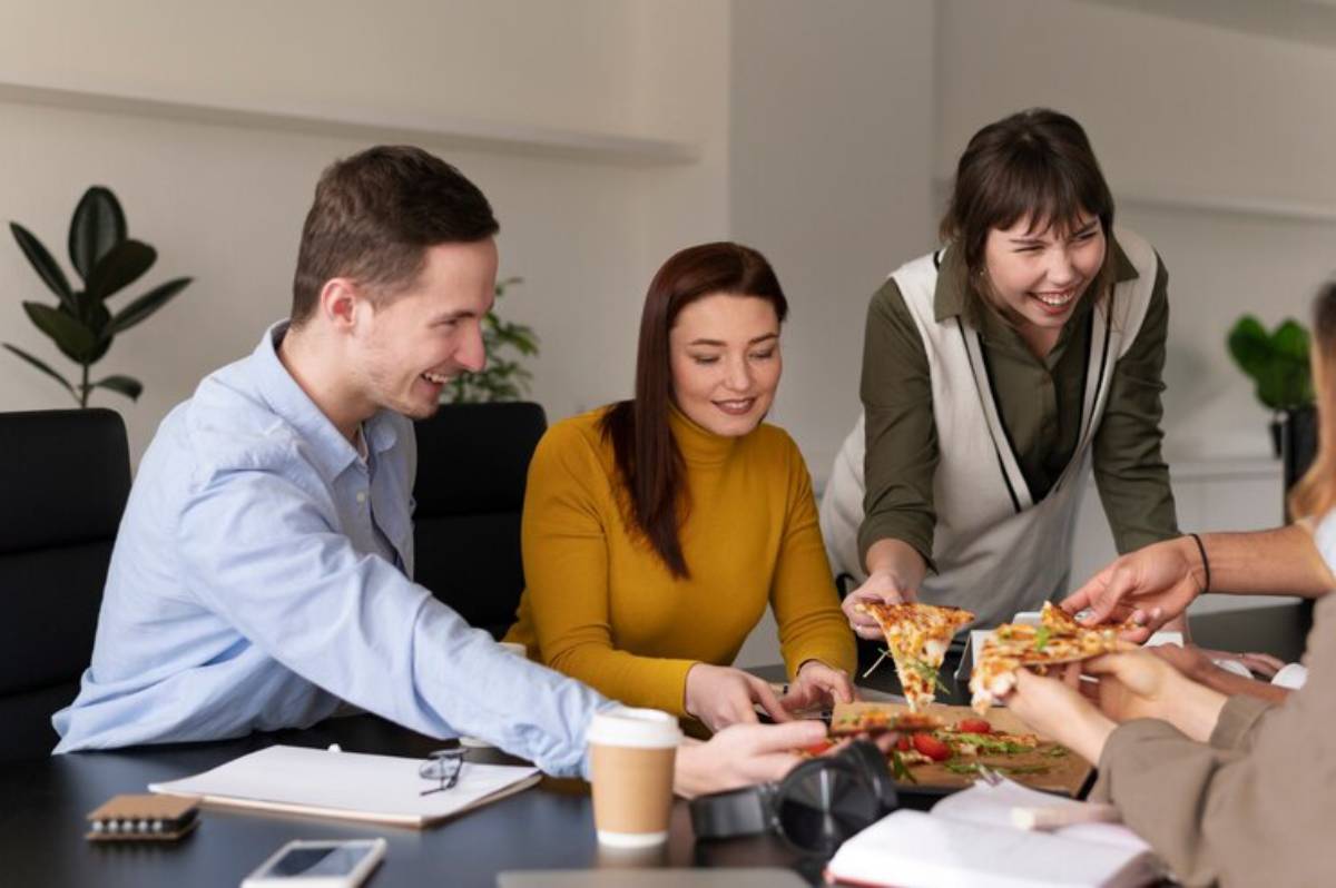 A group of people sharing pizza in a modern office setting, with hands reaching out for slices and a coffee cup on the table.