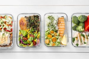 Four meal prep containers filled with colorful, healthy meals: salads, grilled fish, vegetables, and grains on a white background.