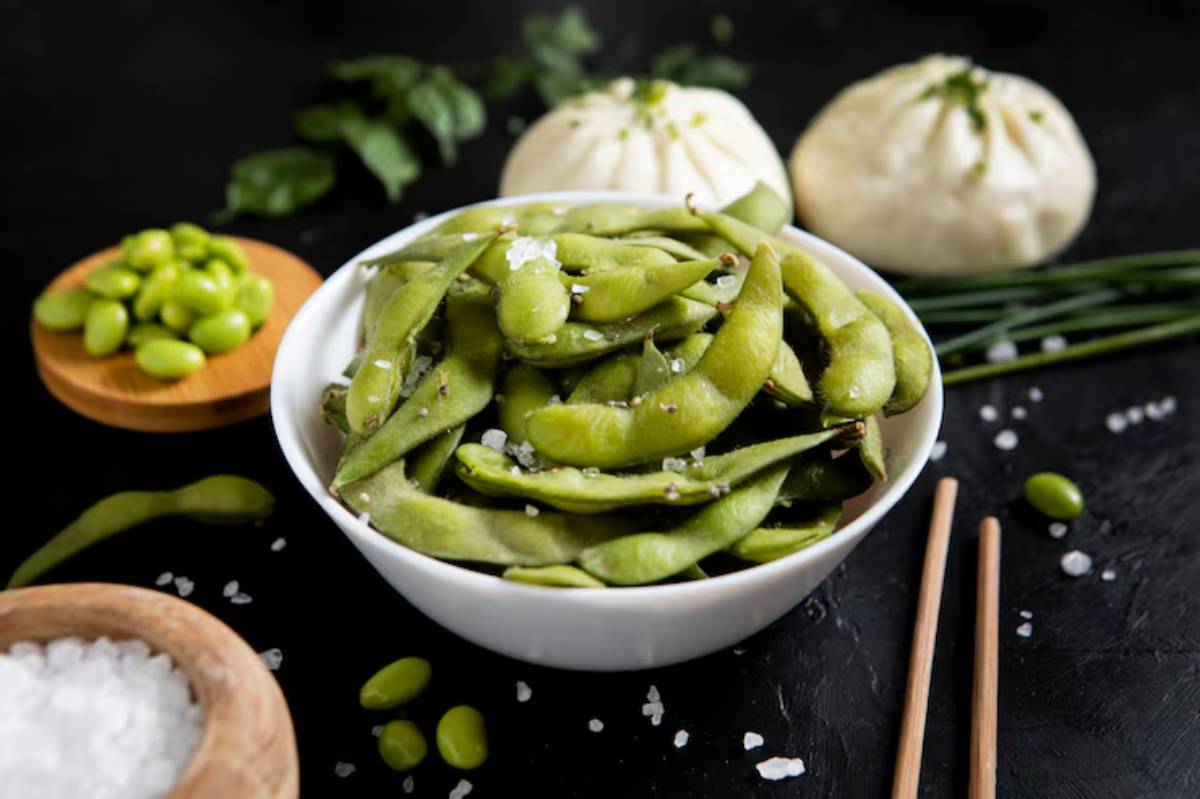 A bowl of salted edamame sits on a dark table, surrounded by fresh green vegetables, bamboo chopsticks, and seasoning salt.