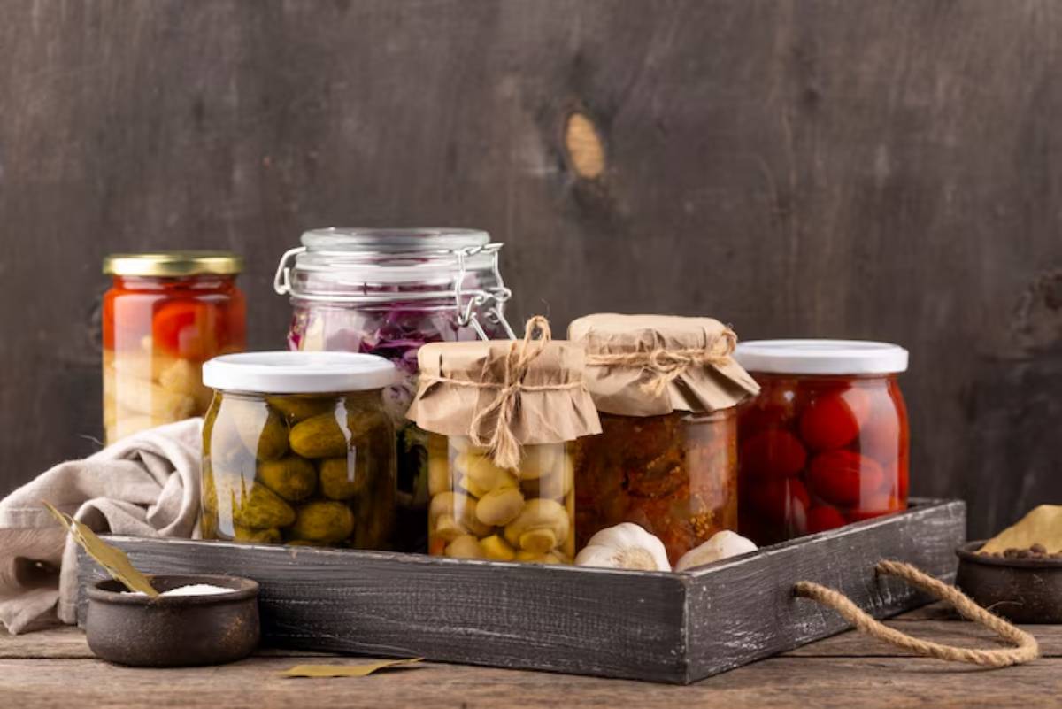 A wooden tray filled with assorted jars of pickled vegetables, olives, and garlic, displayed against a rustic background.