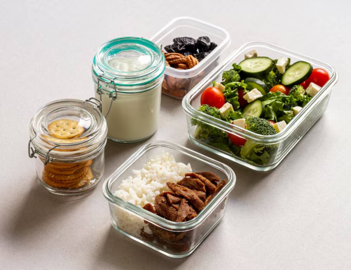 A meal prep arrangement featuring a salad, rice with meat, milk, crackers, and nuts in glass containers on a light surface.