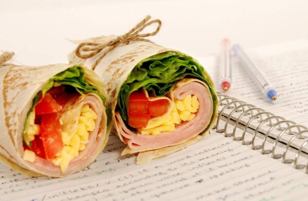 Two neatly wrapped sandwich rolls with lettuce, tomato, ham, and egg, placed next to a notebook and pens.