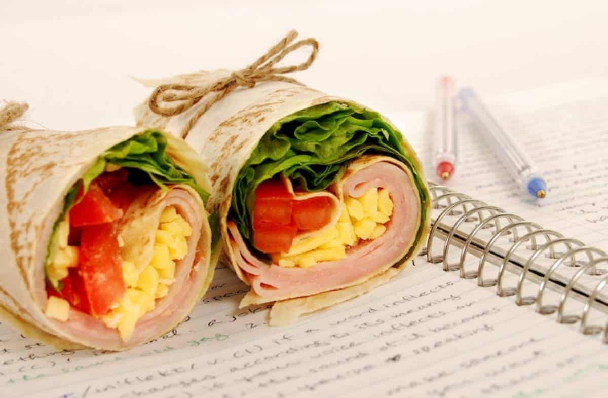 Two neatly wrapped sandwich rolls with lettuce, tomato, ham, and egg, placed next to a notebook and pens.