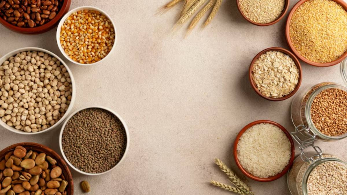 A variety of grains and legumes displayed in bowls on a neutral background, featuring rice, oats, lentils, and wheat stalks.
