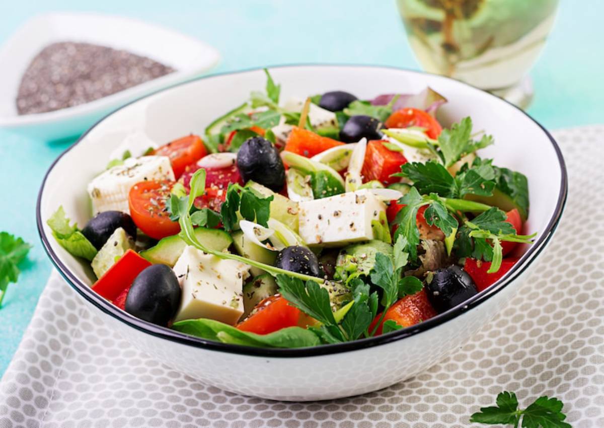 A vibrant salad with mixed greens, tomatoes, cucumbers, olives, feta cheese, and herbs, served in a white bowl with a decorative background.