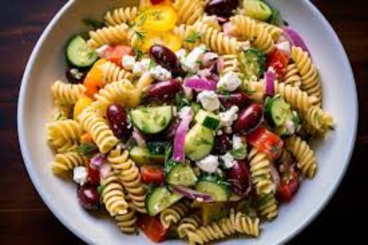 A colorful pasta salad with rotini, cherry tomatoes, cucumbers, red onions, kidney beans, herbs, and feta cheese in a large bowl.