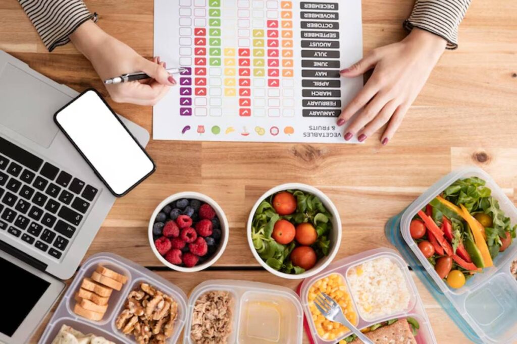 Top view of a workspace with a laptop, phone, and colorful meal prep containers filled with fruits and vegetables, alongside a chart.