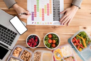 Top view of a workspace with a laptop, phone, and colorful meal prep containers filled with fruits and vegetables, alongside a chart.