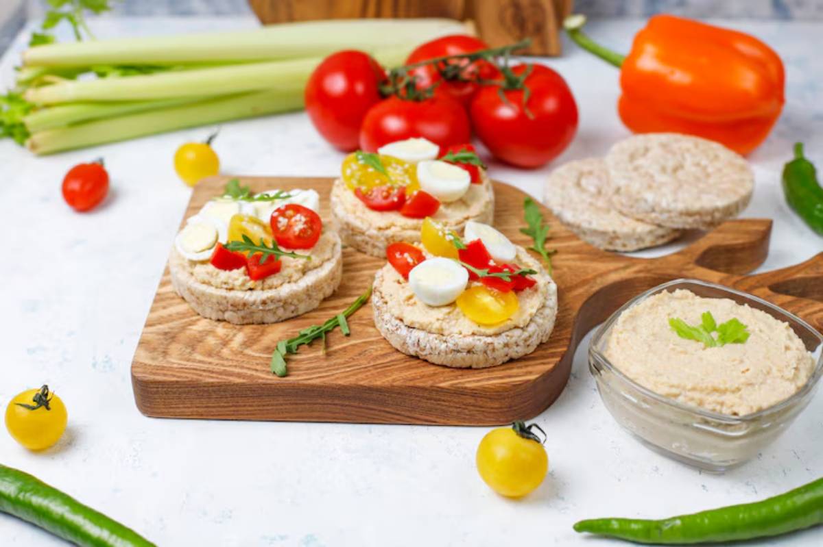 A wooden board with rice cakes topped with cherry tomatoes, cheese, and arugula, accompanied by a bowl of creamy dip and fresh vegetables.