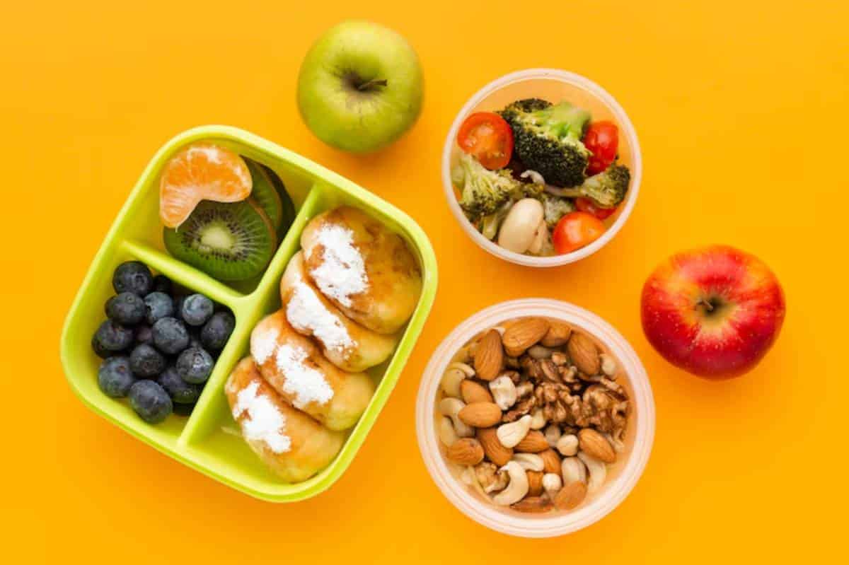 A colorful lunch featuring a green container with fruit, a salad bowl, and a snack of mixed nuts, set against a bright orange background.