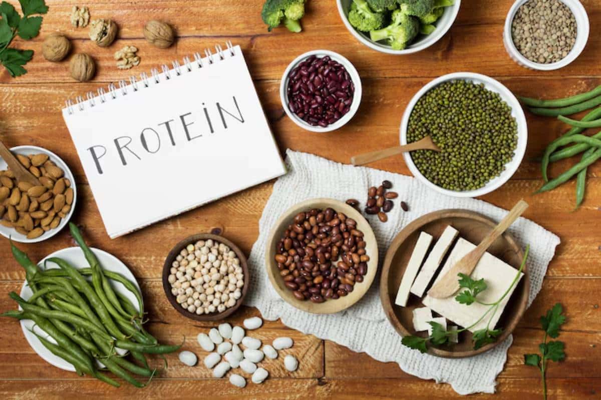 A variety of plant-based protein sources, including beans, tofu, almonds, and broccoli, arranged on a wooden table with a notepad labeled