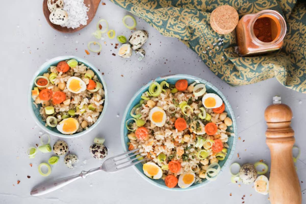 A colorful grain salad with carrots, leeks, and quail eggs, served in two bowls with a fork, spices, and salt on a textured background.