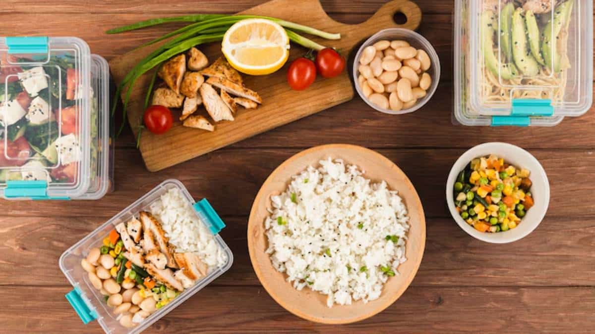 A variety of meal prep containers with chicken, rice, vegetables, salad, and fresh ingredients arranged on a wooden surface.
