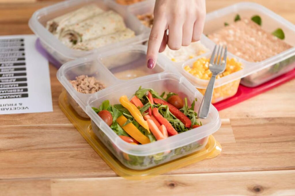 A hand points to a container filled with a colorful salad of greens, cherry tomatoes, and sliced bell peppers on a wooden table.