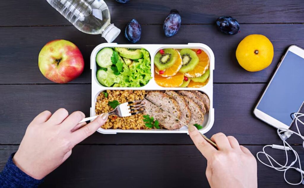A hand holds a fork over a healthy lunchbox with grains, meat, salad, and fruit, accompanied by an apple, a water bottle, and a smartphone.