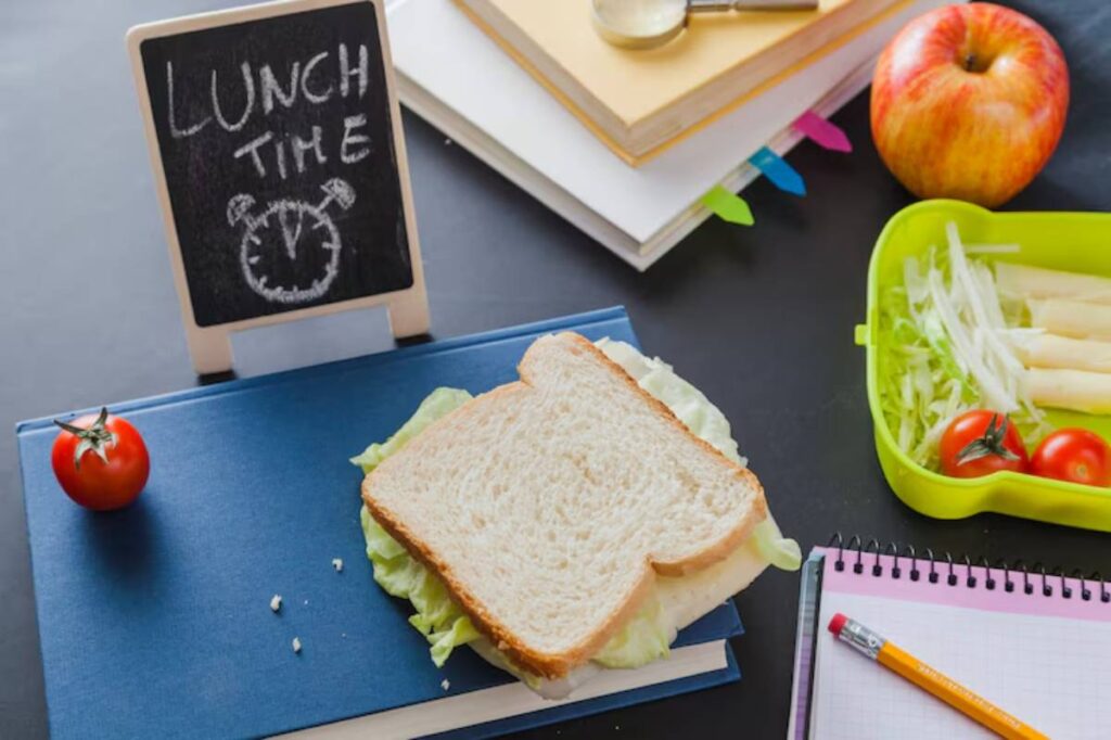 A sandwich on a book, surrounded by a tomato, an apple, a lunchbox with veggies, and a chalkboard reading "LUNCH TIME."